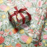 A wrapped gift box tied with a dark red ribbon sits on top of matching wrapping paper printed with a whimsical botanical and bird pattern. The design features small birds, pink flowers, yellow and red botanical elements, and ornaments on a pale sage green background.