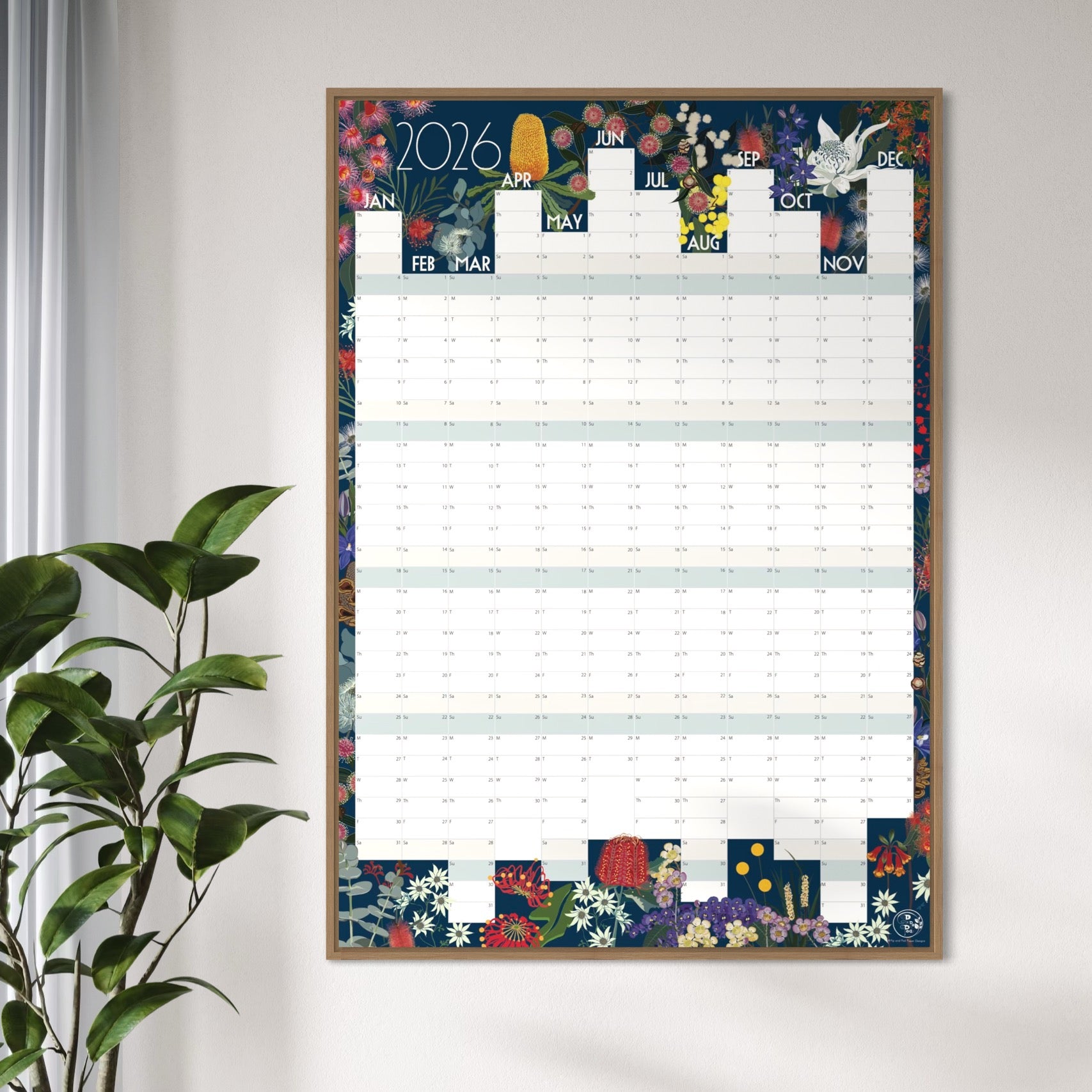 A framed 2026 wall calendar hanging on a light gray wall, styled next to a green potted plant. The calendar features a colorful floral border and illustrated monthly grids.