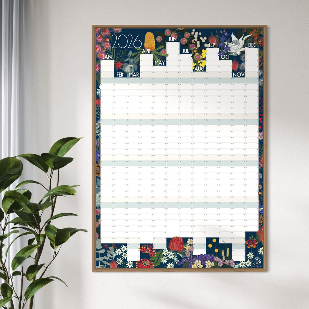 A framed 2026 wall calendar hanging on a light gray wall, styled next to a green potted plant. The calendar features a colorful floral border and illustrated monthly grids.