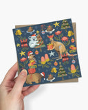 A hand holds a square Christmas-themed card featuring a navy blue background with illustrated kangaroo, koala, kangaroo tail, Christmas tree, ornaments, gifts, and stars, layered over a brown envelope against a light gray background.