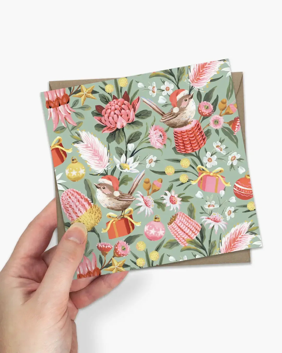 A hand holds a square greeting card with a festive Australian-themed pattern featuring birds, protea flowers, kangaroo rattles, and Christmas ornaments on a sage green background, with a brown envelope partially visible behind it.