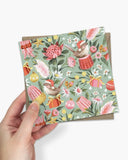A hand holds a square greeting card with a festive Australian-themed pattern featuring birds, protea flowers, kangaroo rattles, and Christmas ornaments on a sage green background, with a brown envelope partially visible behind it.