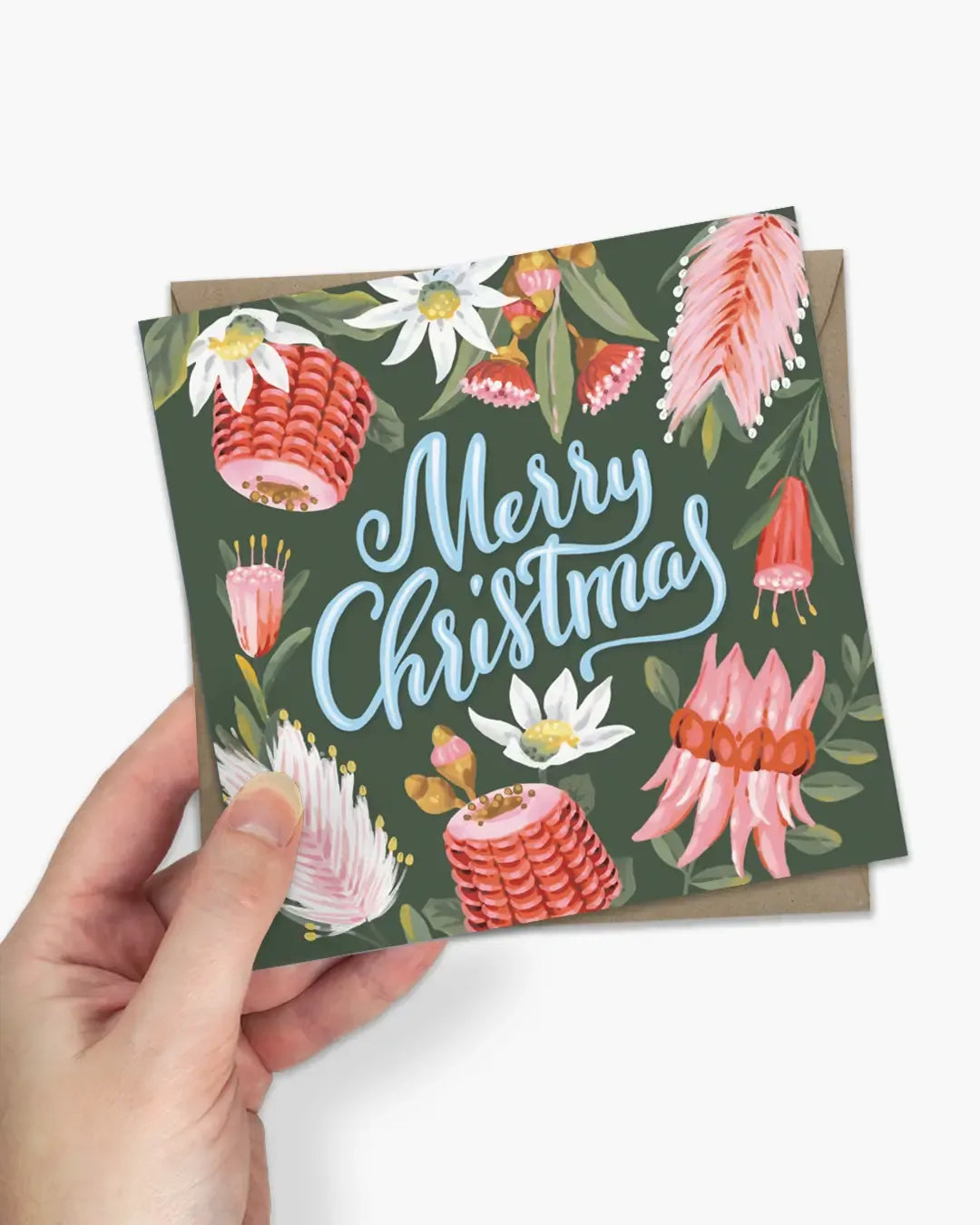 A hand holds a square Christmas greeting card featuring a dark green background with watercolor-style pink and white flowers, green leaves, and the words 'Merry Christmas' in light blue script. A brown envelope is partially visible behind the card against a clean white background.