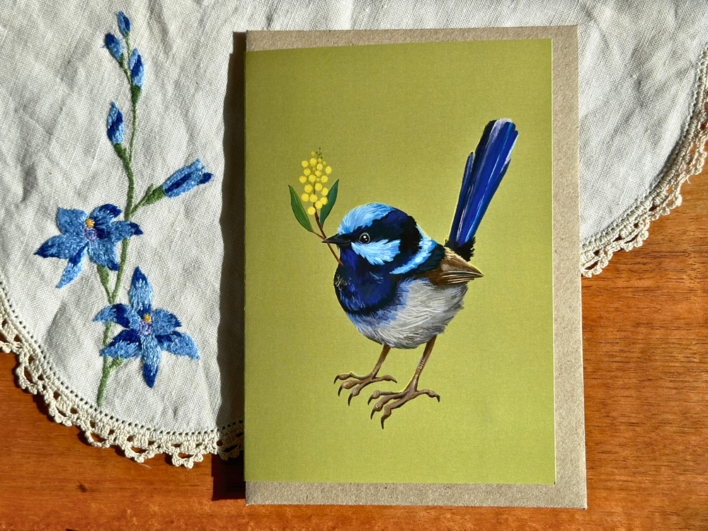 A greeting card featuring a detailed illustration of a blue and white fairy wren holding a yellow flower, displayed on a wooden table next to a white embroidered cloth.