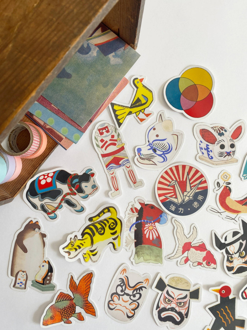 A collection of colorful, die-cut stickers featuring Japanese cultural motifs, animals, and faces, arranged on a white surface next to a wooden box.
