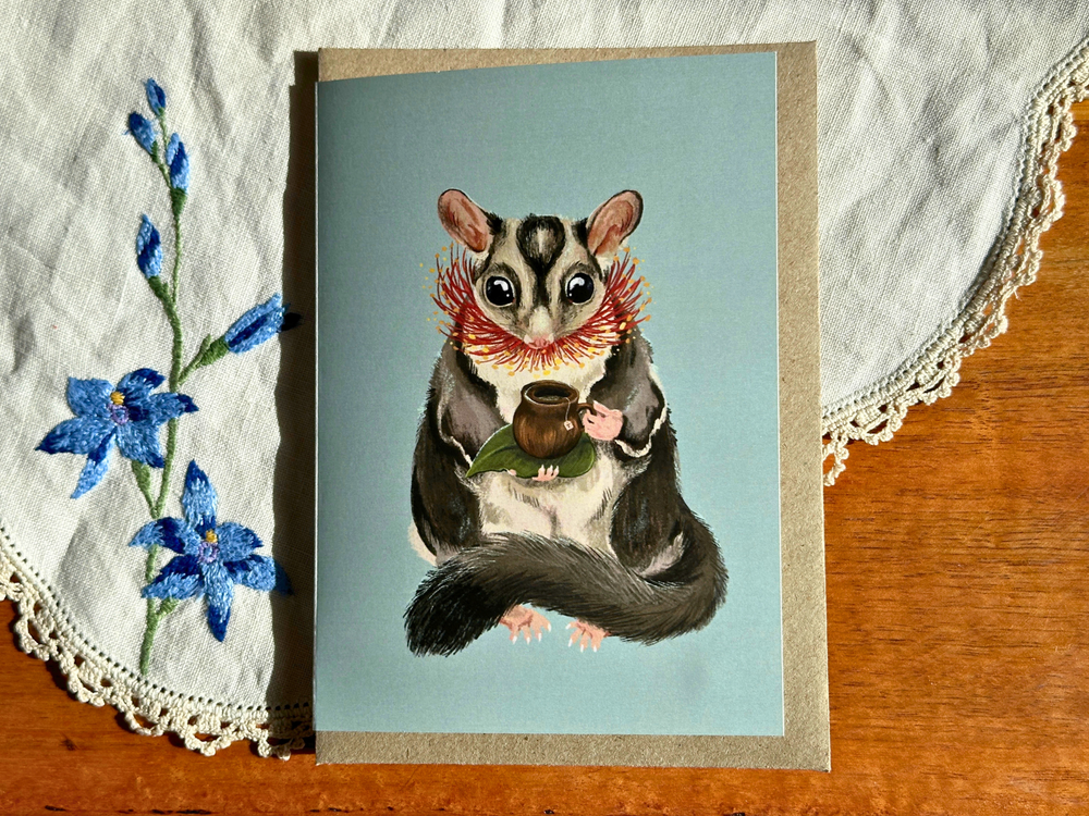 Greeting card with a sugar glider illustration on a blue background, placed on a wooden surface with lace and flowers.