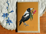 A card featuring a detailed illustration of a magpie holding red flowers, displayed on a wooden surface next to a white embroidered cloth.