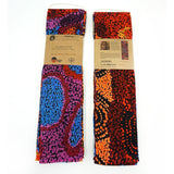 Two folded cotton scarves with brown paper bands are displayed side-by-side against a white background. The scarf on the left features a blue, purple, and red abstract pattern, while the scarf on the right features an orange, red, and black dot pattern.