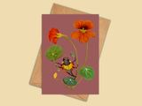 A botanical art print card featuring orange nasturtiums and a yellow-and-black ladybug on a dusty rose background, shown with a kraft paper envelope on a warm beige backdrop.