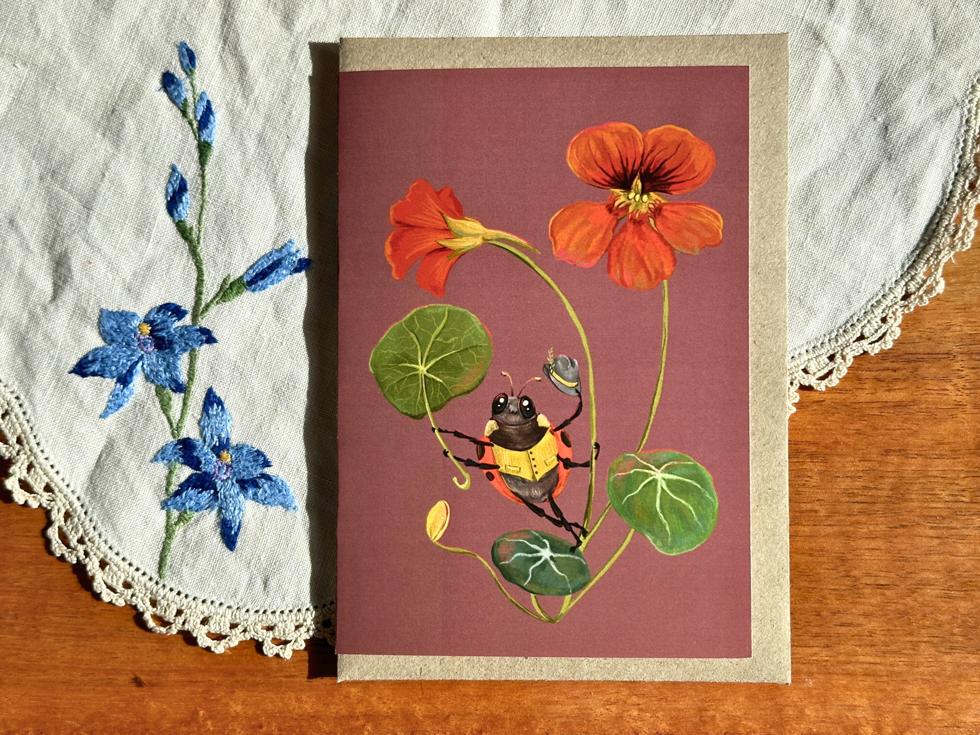 A rectangular art print or card featuring a botanical illustration of orange flowers, green leaves, and a yellow-and-black bee on a dusty mauve background, placed on a wooden surface with a white embroidered cloth underneath.