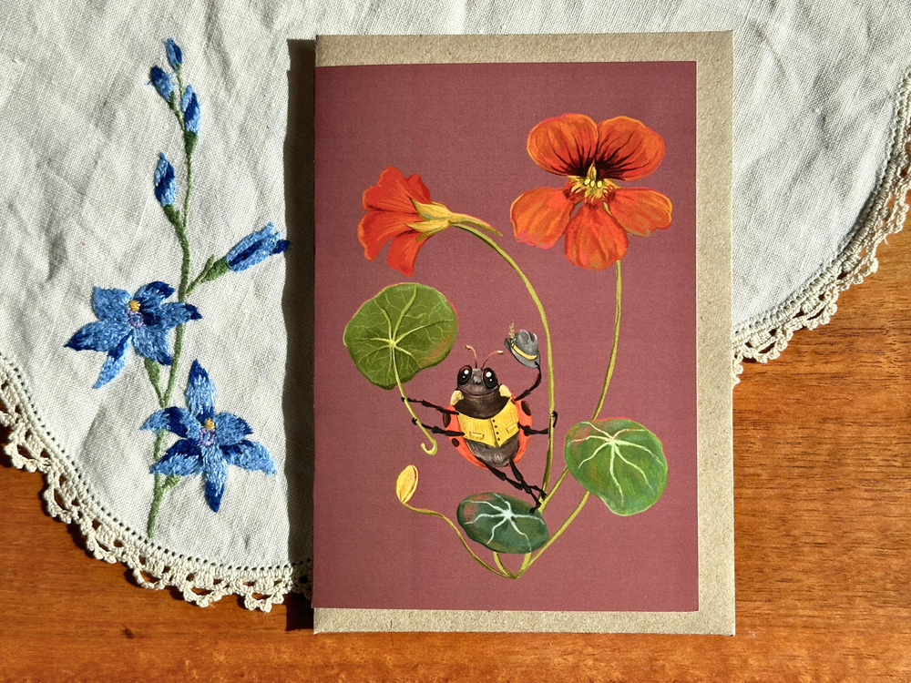 A rectangular art print or card featuring a botanical illustration of orange flowers, green leaves, and a yellow-and-black bee on a dusty mauve background, placed on a wooden surface with a white embroidered cloth underneath.
