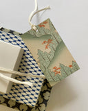 A rectangular gift tag with a Japanese-style landscape print featuring green trees and orange blossoms, tied with a white string to a wrapped gift box on a plain light background.