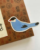 A die-cut sticker of a stylized blue bird with a black head, white beak and outline, and red beak tip, placed on top of a brown patterned paper envelope.